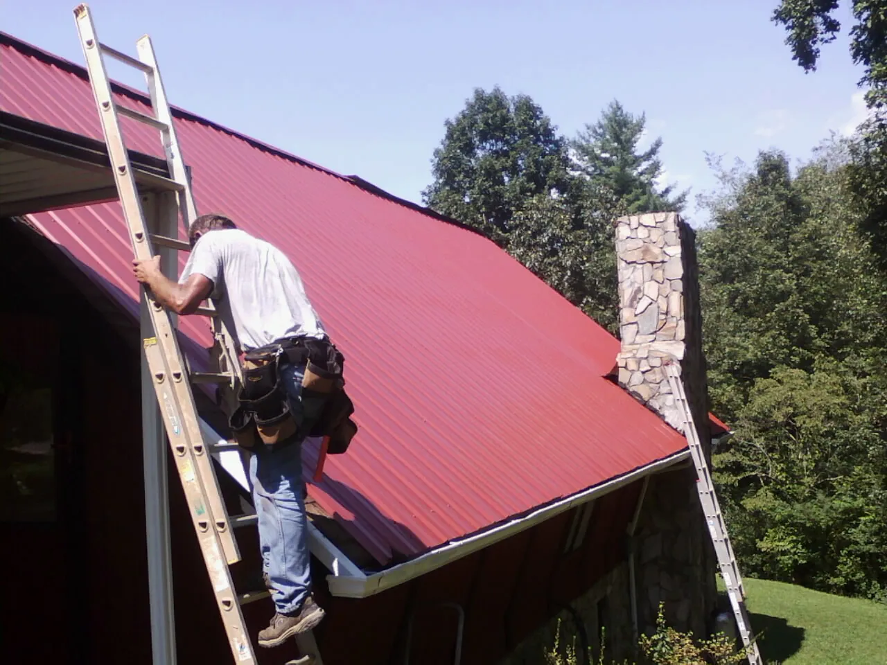 Roofer on a ladder beside a red metal roof and stone chimney — Soffit & Fascia Repair by Roofing Brothers in Gerlach